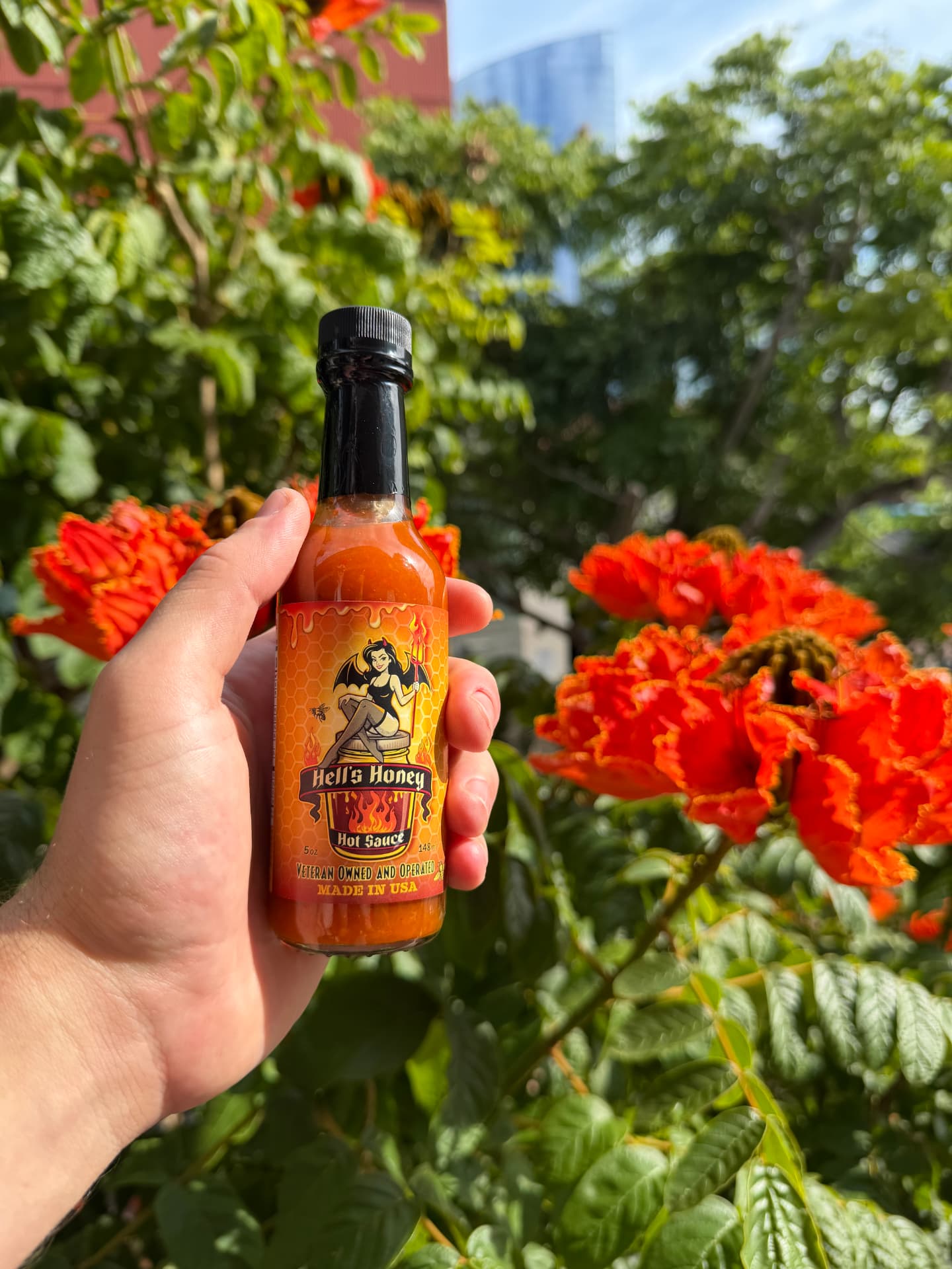 A hand holding a bottle of Hell's Honey Hot Sauce in front of bright orange flowers
